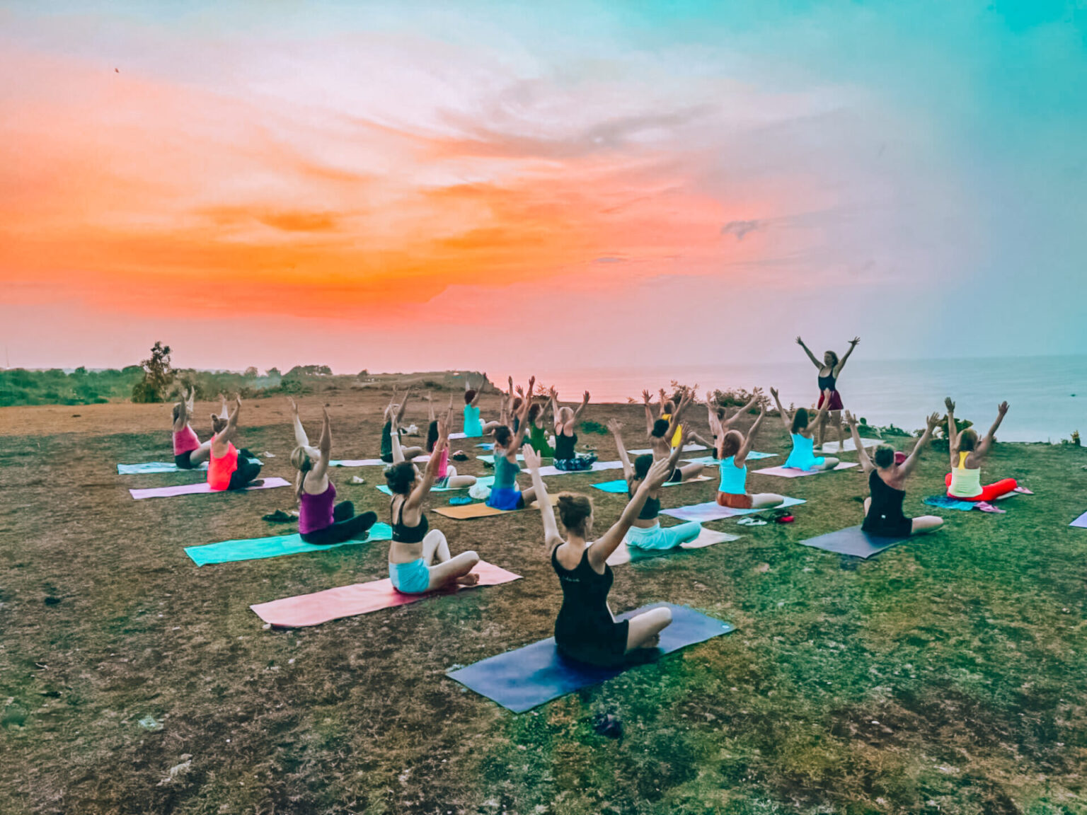 Judita vede jógovou praxi na písečné pláži u tyrkysového oceánu na Bali, obklopena účastnicemi retreatu zaujímajícími pozice v ranním slunci, s palmami a tradicí posvátné transformace.