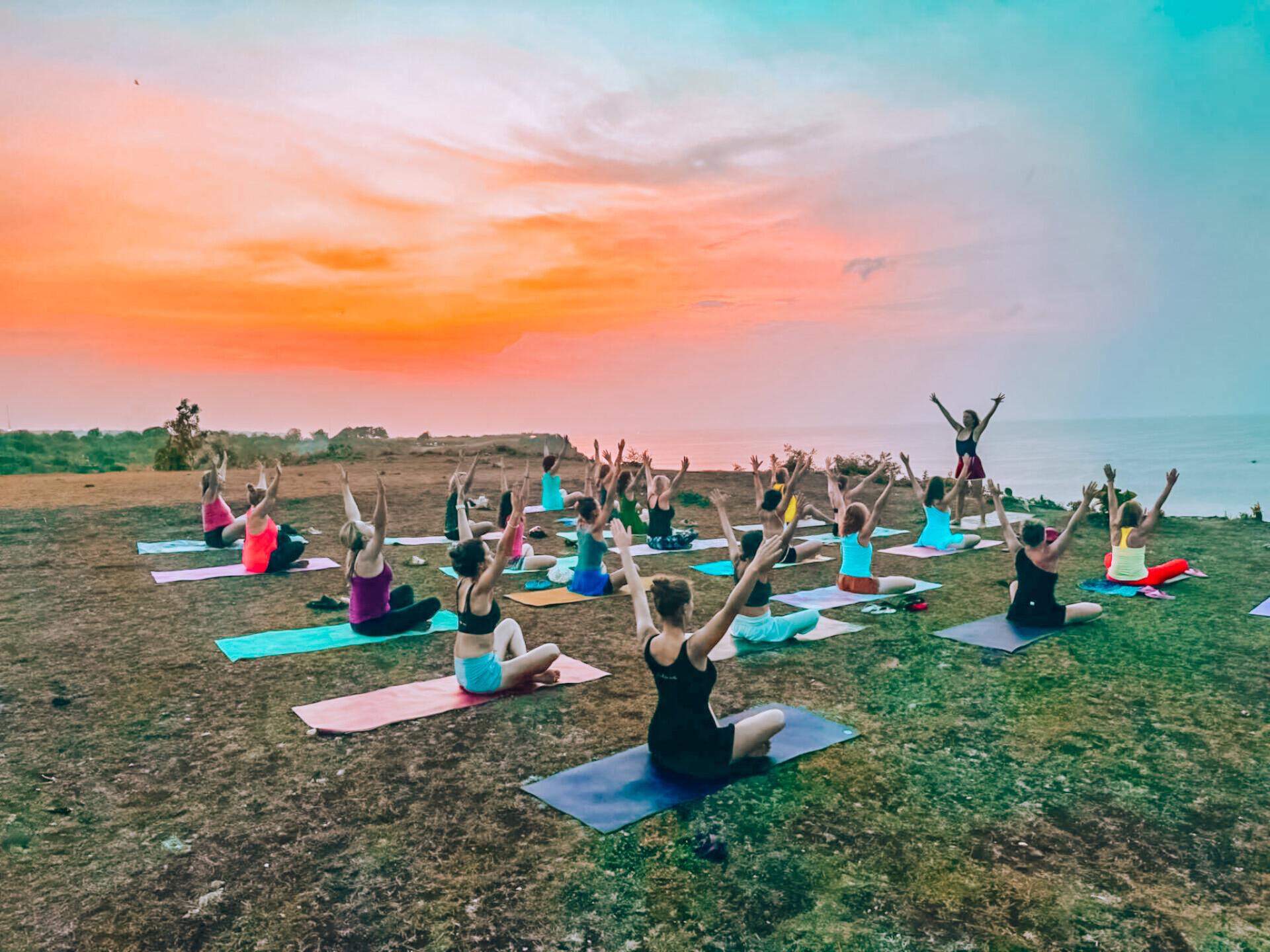 Judita vede jógovou praxi na&nbsp;písečné pláži u&nbsp;tyrkysového oceánu na&nbsp;Bali, obklopena účastnicemi retreatu zaujímajícími pozice v&nbsp;ranním slunci, s&nbsp;palmami a&nbsp;tradicí posvátné transformace.